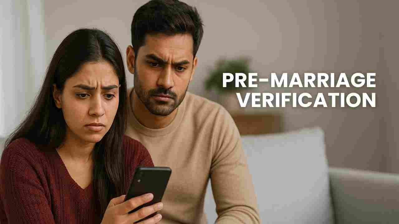 A young South Asian couple sitting together on a sofa, both looking seriously at a smartphone, reflecting concern and investigation — concept of pre-marriage verification and background checks.